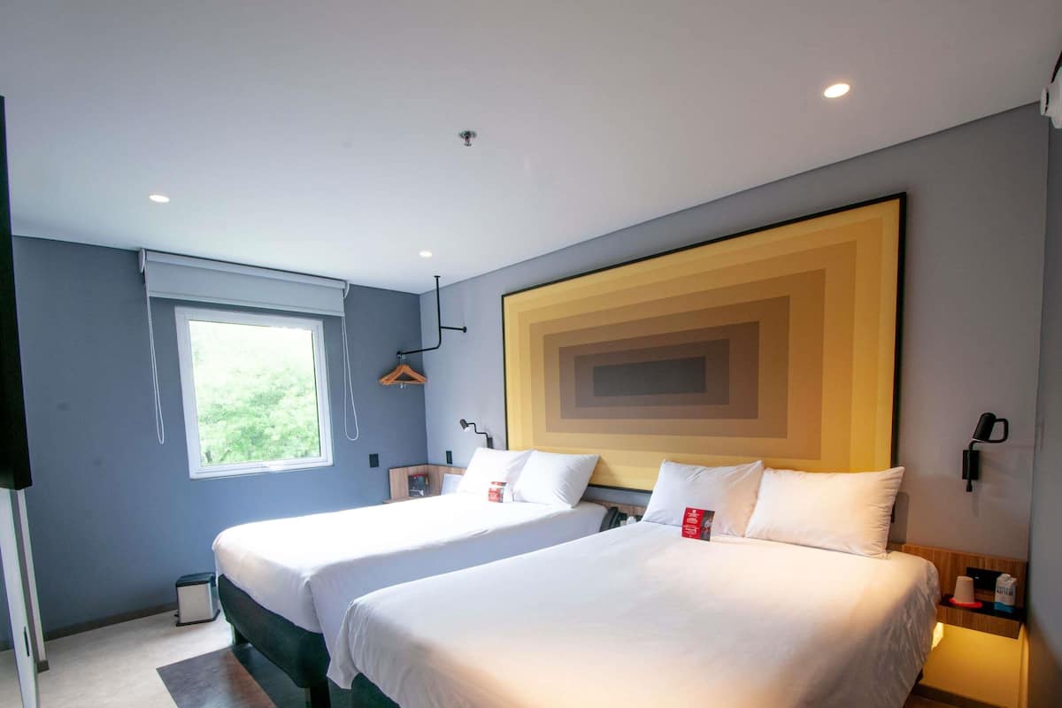 Habitación de hotel con dos camas, un ventanal grande, una lámpara de pared y un cubo de basura pequeño.