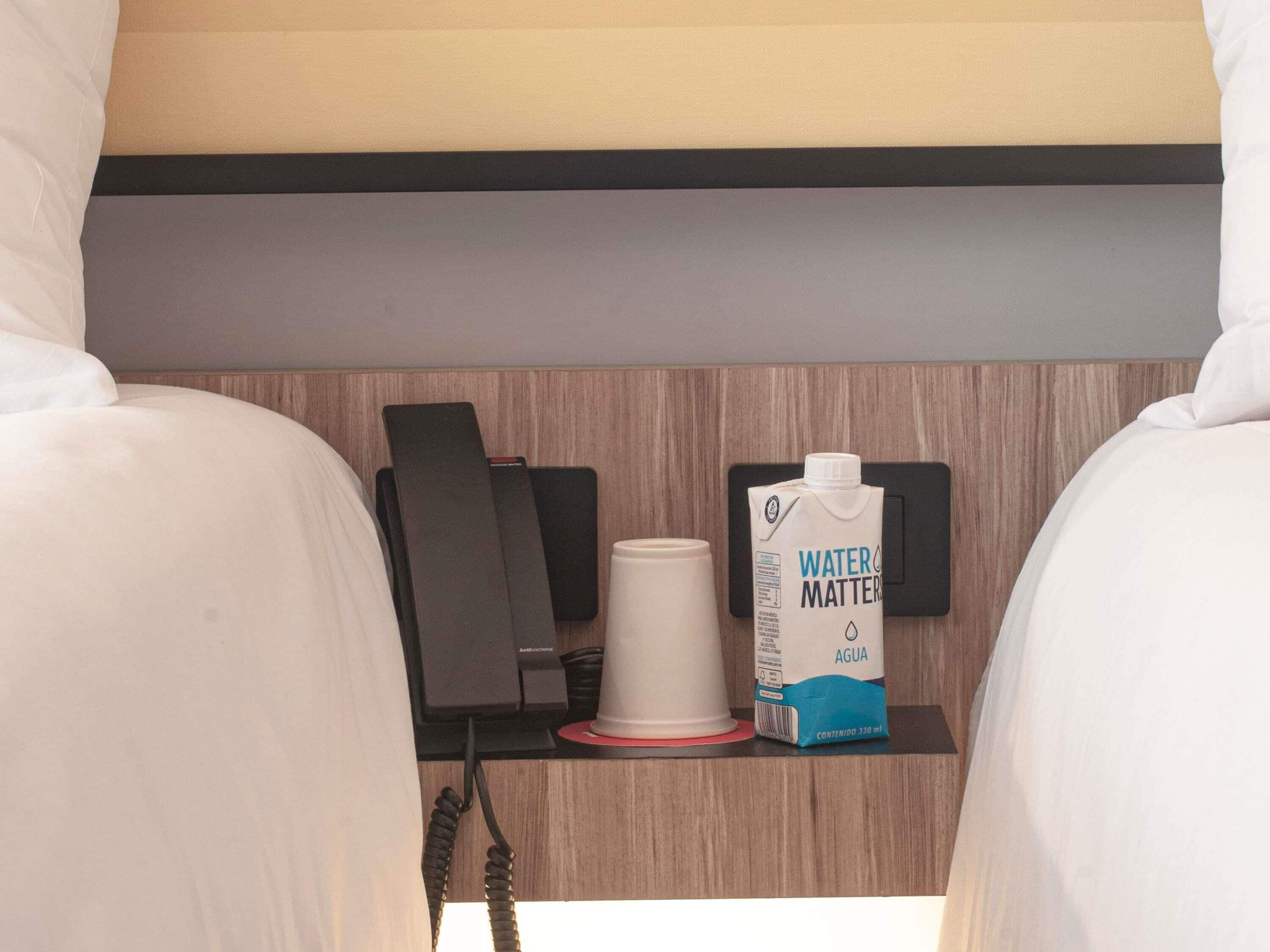 Una habitación de hotel con cama, mesita de noche, teléfono, taza y botella de agua.