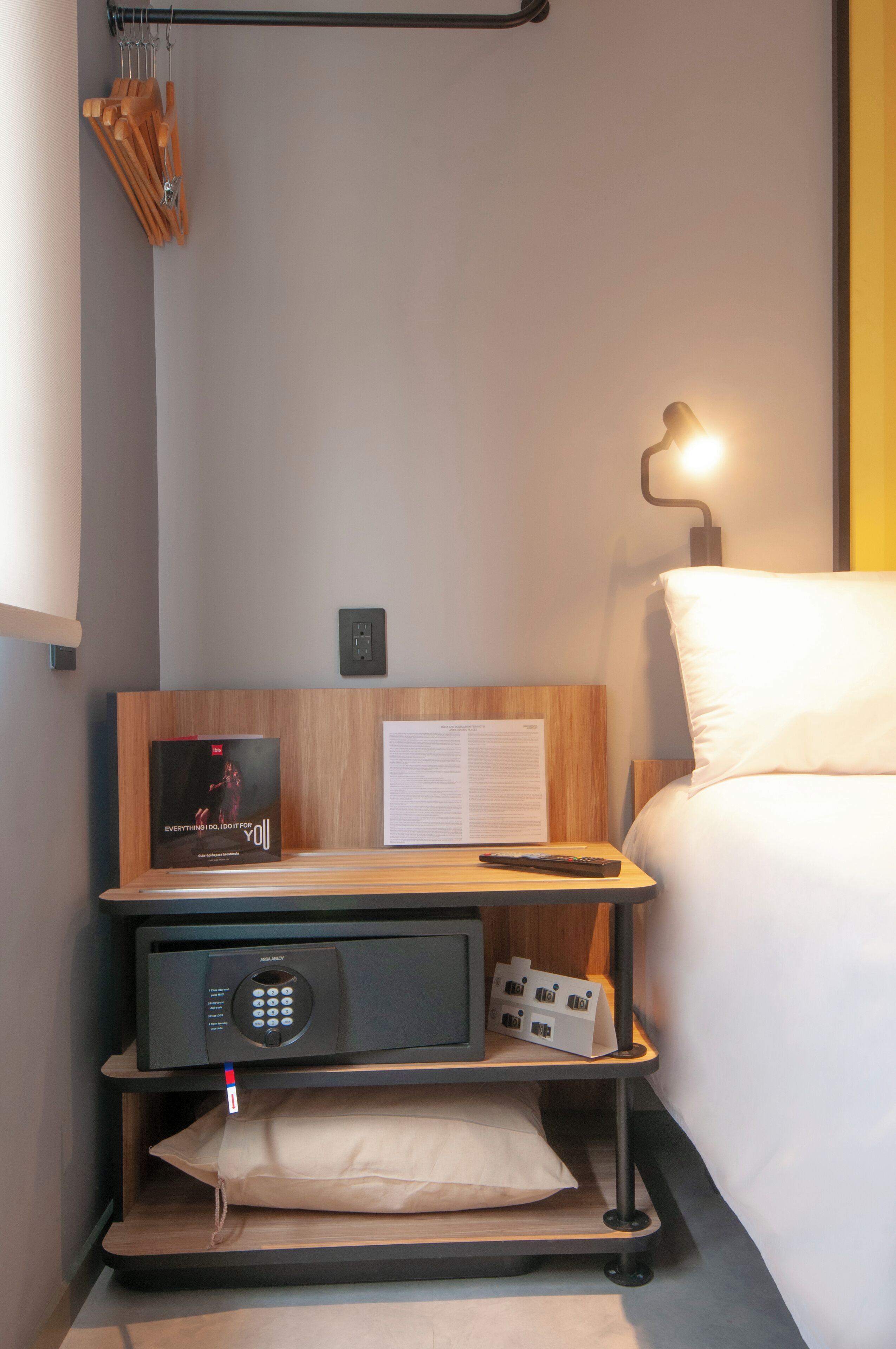 Una habitación de hotel ordenada con una mesita de noche de madera, un control remoto, un reloj digital y una lámpara de noche.