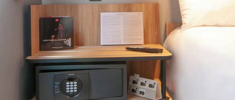 Una habitación de hotel ordenada con una mesita de noche de madera, un control remoto, un reloj digital y una lámpara de noche.