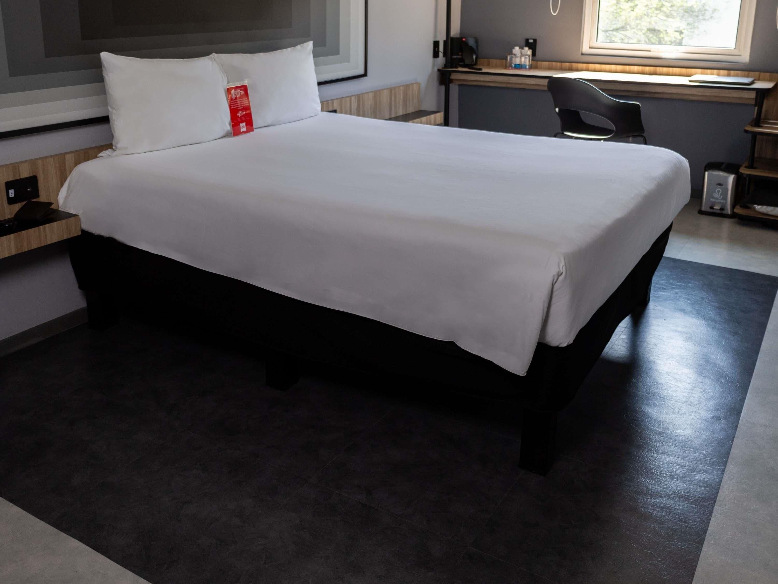 Een netjes opgemaakt bed in een hotelkamer met een bureau, een stoel, een raam en een klein nachtkastje.