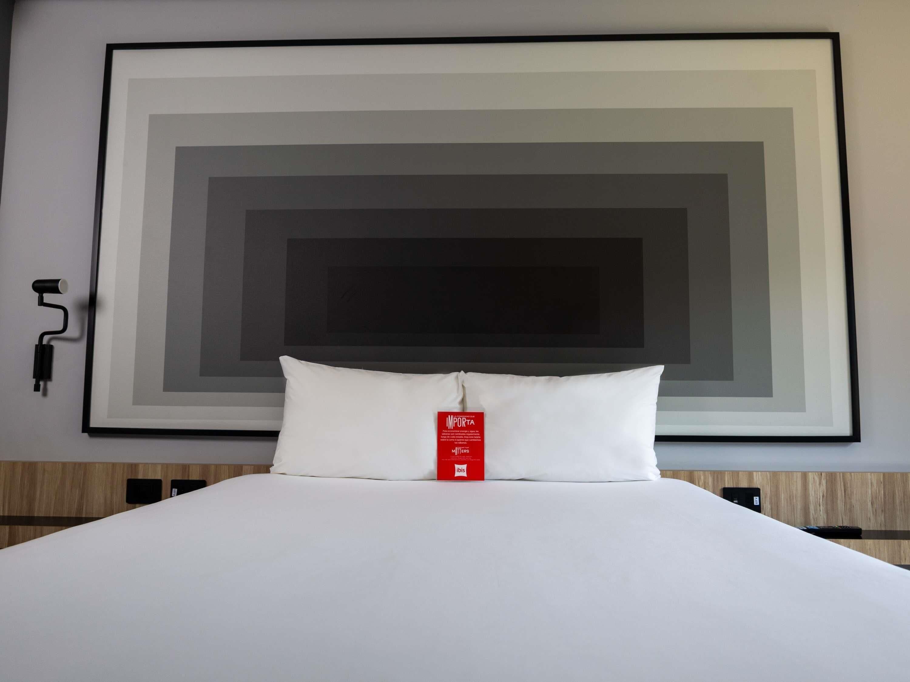 A hotel room with a large framed artwork on the wall, a white bed with a pillow, and a red tag on the pillow.