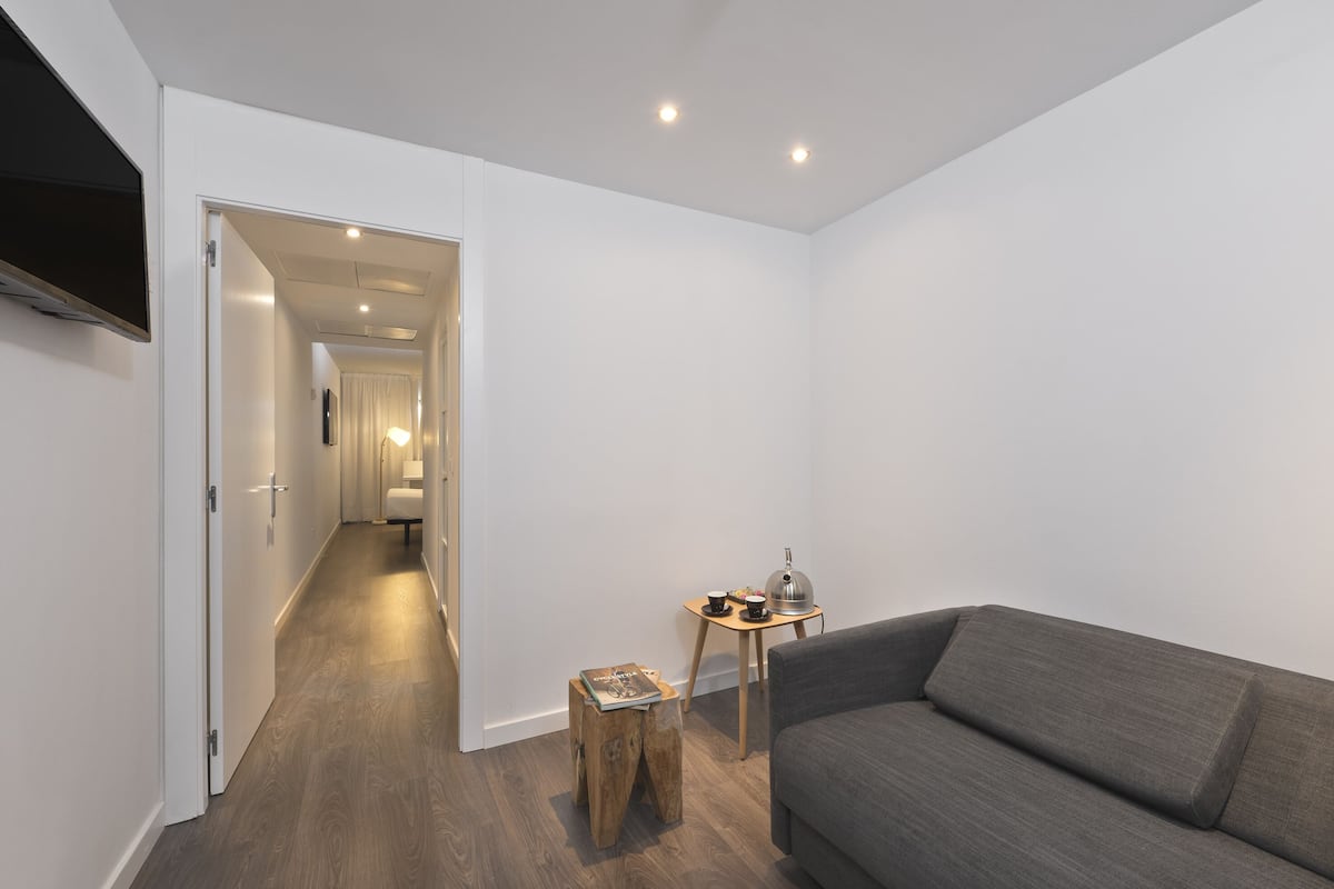 Ein modernes Wohnzimmer mit einem grauen Sofa, einem Holztisch und einem kleinen Beistelltisch mit Lampe. Ein Flur führt zu einem Badezimmer mit Toilette und Dusche.