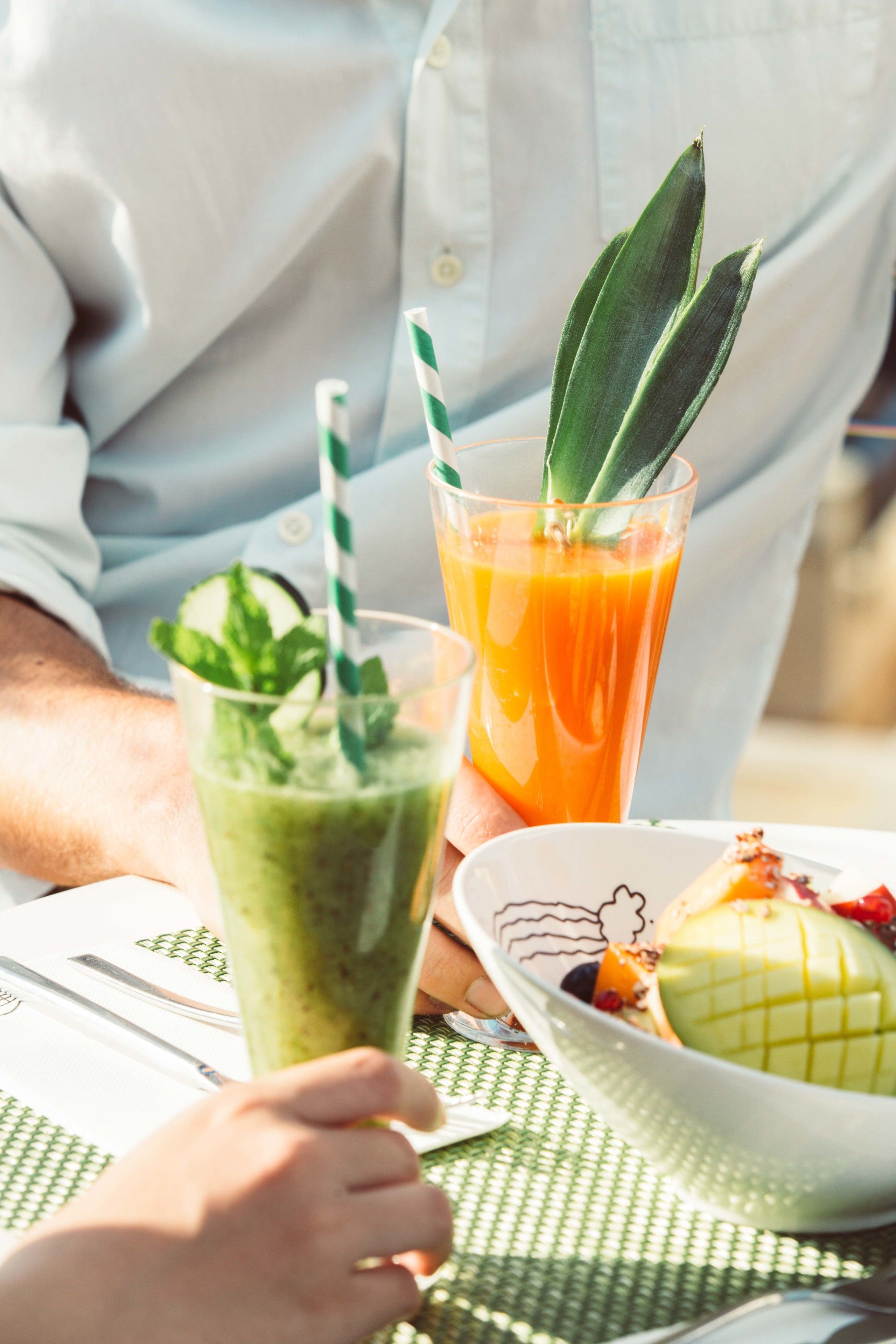 A person holding a green smoothie with a mint garnish, a glass of orange juice, and a bowl of fruit on a table.