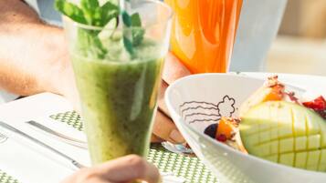 Una persona sostiene un batido verde con adorno de menta, un vaso de jugo de naranja y un bol de fruta sobre una mesa.