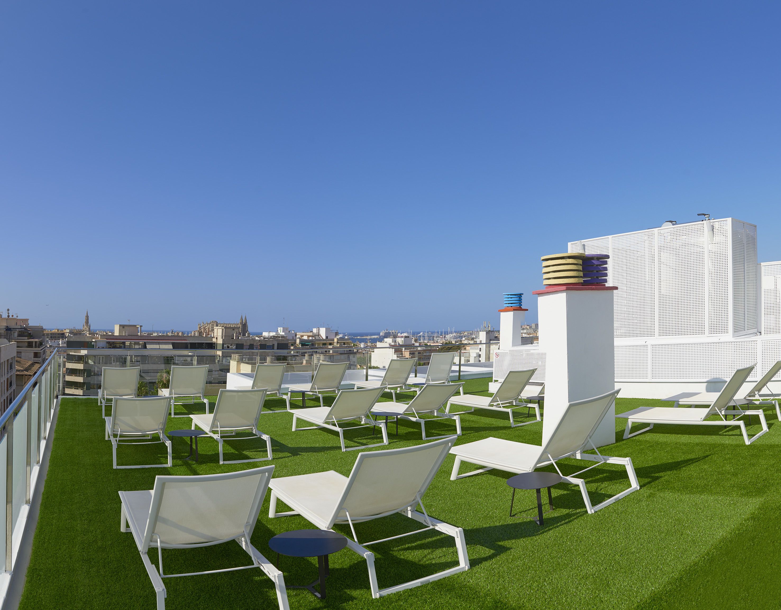 Terraza en la azotea con sillas de descanso blancas y césped artificial, con vistas a la ciudad y al mar.