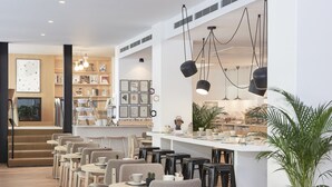 A modern cafe with a long counter, multiple tables, and chairs. There are hanging pendant lights above the counter and a staircase leading to an upper level. - INNSiDE by Meliá Palma Center (Palma de Mallorca)