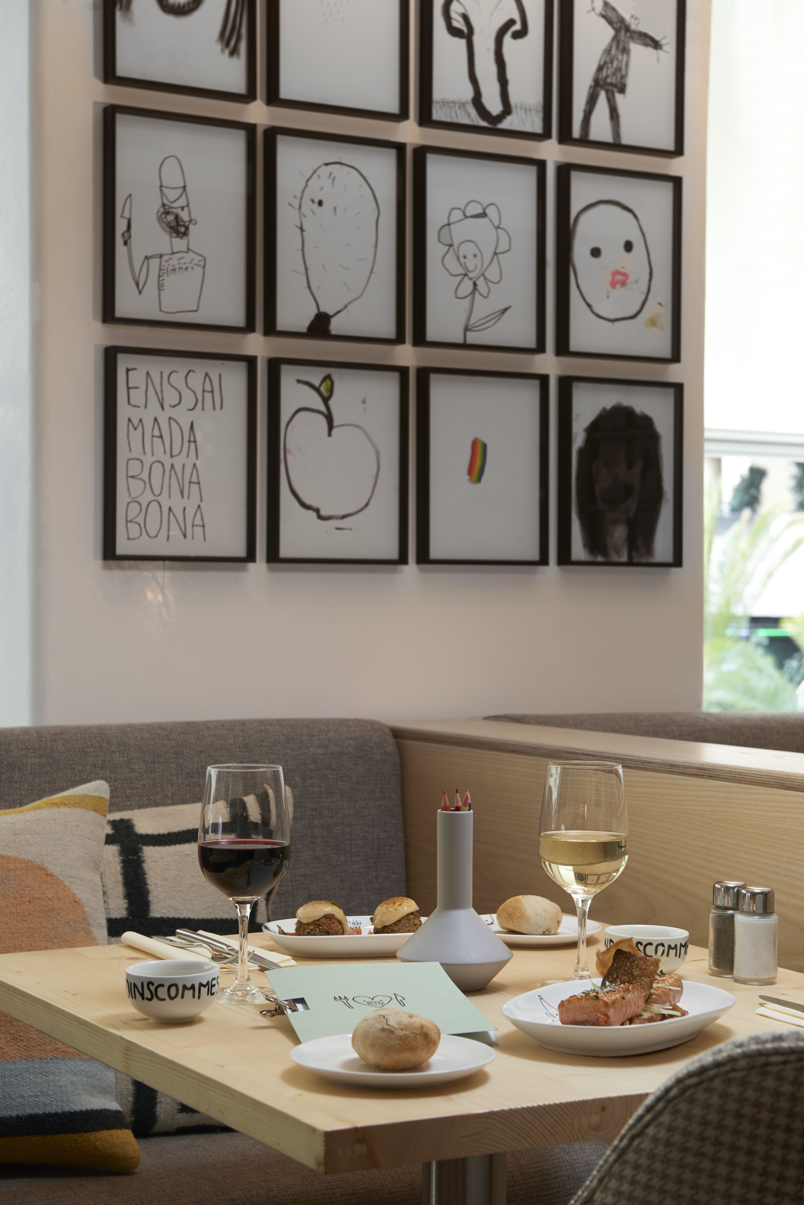Un comedor con una mesa dispuesta para dos personas, que incluye platos de comida, copas de vino y una vela. En la pared detrás de la mesa se exhiben obras de arte enmarcadas.