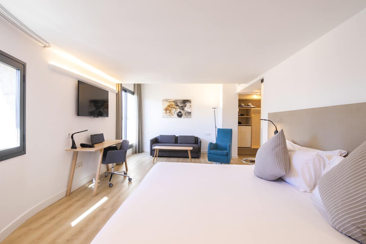 Ein Hotelzimmer mit einem großen Bett, einem Schreibtisch mit Computer, einer Couch und einem an der Wand befestigten Fernseher.