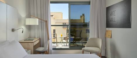 A hotel room with a bed, a desk with a lamp, a chair, a small table with a glass of orange juice, and a view of the outside through a glass door.