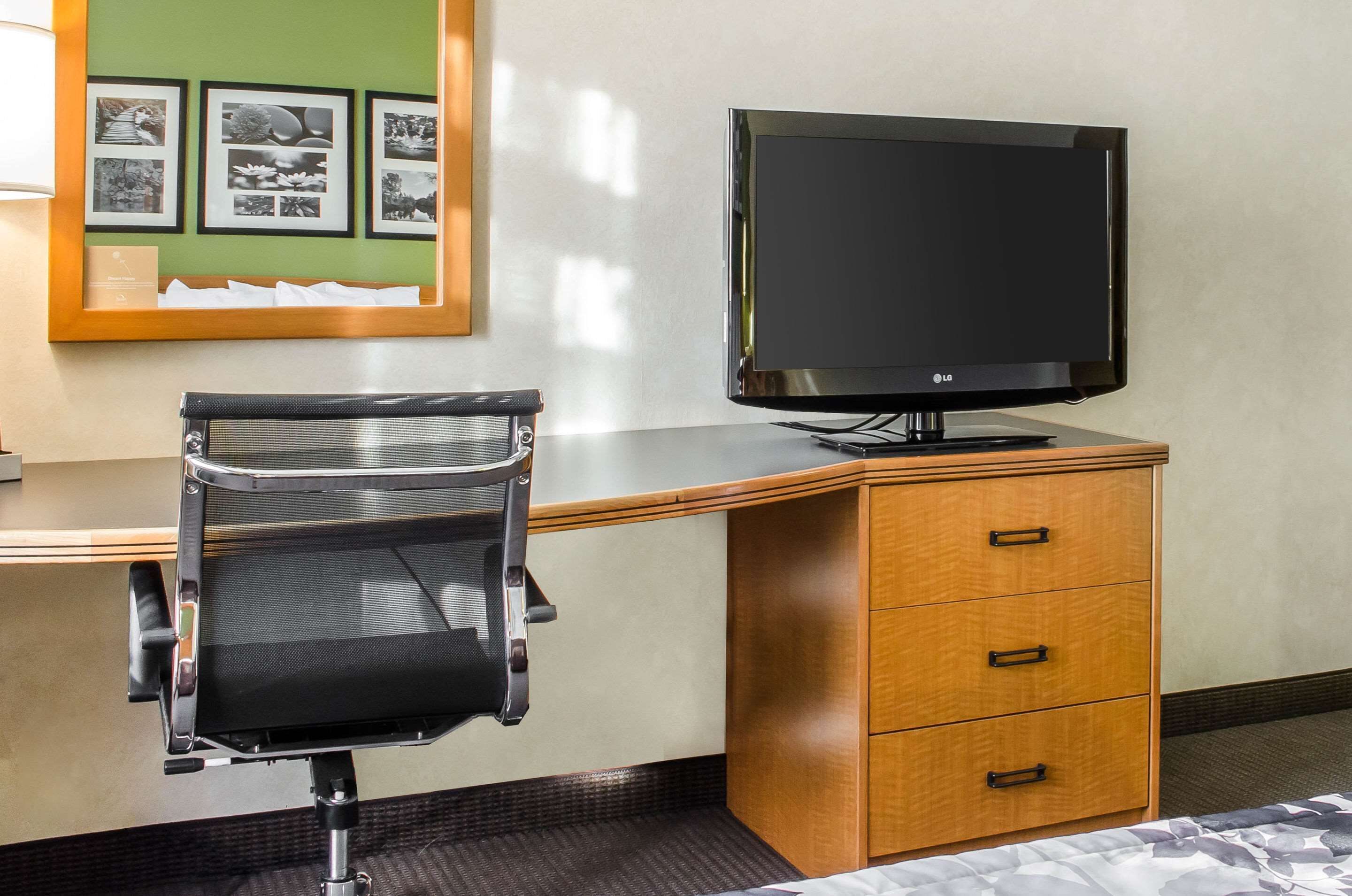 Habitación de hotel con un escritorio de madera, una silla de oficina negra, un televisor de pantalla plana y un expositor de fotos en la pared.