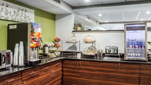 A self-service breakfast area with a juice dispenser, coffee maker, and a refrigerator with beverages.