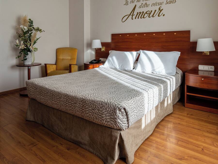 A hotel room with a large bed, a wooden headboard, bedside tables with lamps, a chair, and a decorative wall piece.