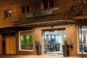 El exterior del Hotel Avenida, con una amplia entrada de vidrio, plantas en macetas y un letrero sobre la entrada.