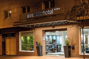 El exterior del Hotel Avenida, con una amplia entrada de vidrio, plantas en macetas y un letrero sobre la entrada.