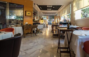 Interior de un restaurante con suelo de mármol, mesas dispuestas con manteles blancos y sillas rojas.