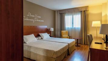 Quarto de hotel com duas camas, uma escrivaninha, uma cadeira, um abajur e janela com cortinas.