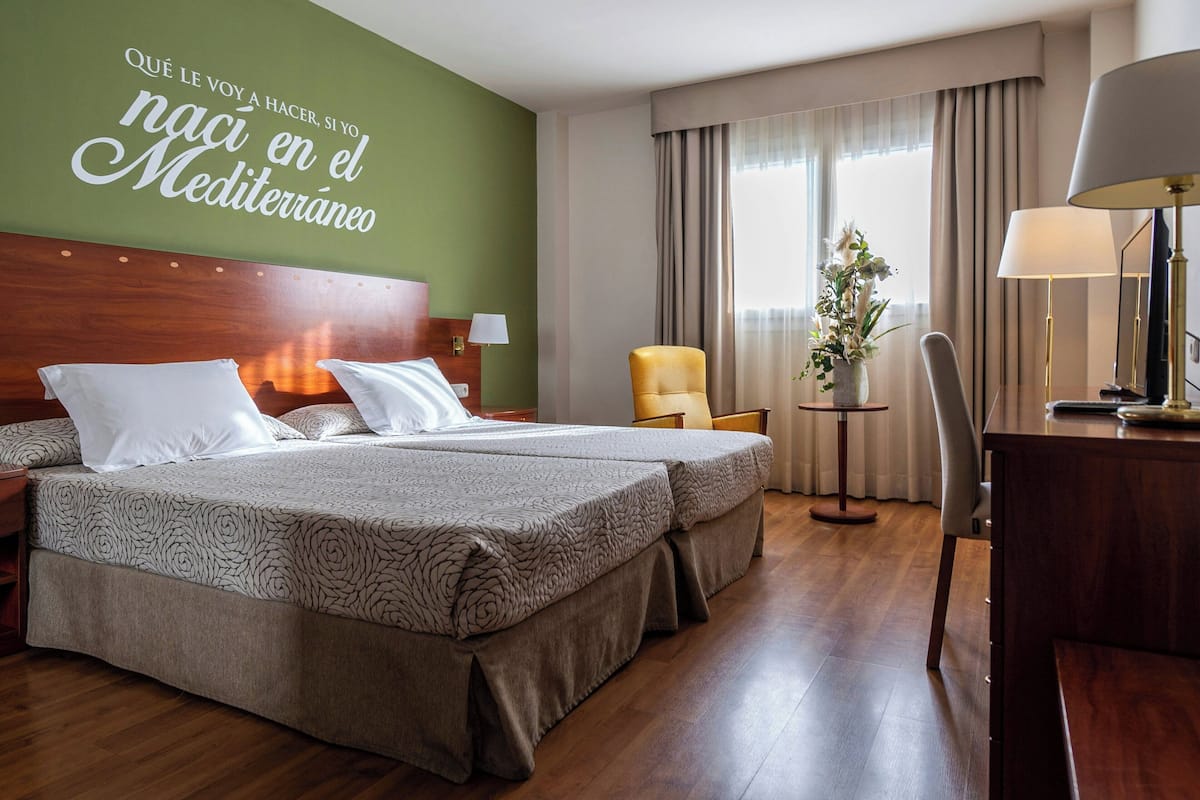 A hotel room with two beds, a desk, a chair, a small table, and a decorative wall with text.