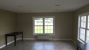 Ein Zimmer mit beigen Wänden, einem großen Fenster, einem dunklen Holztisch und einem Fliesenboden.