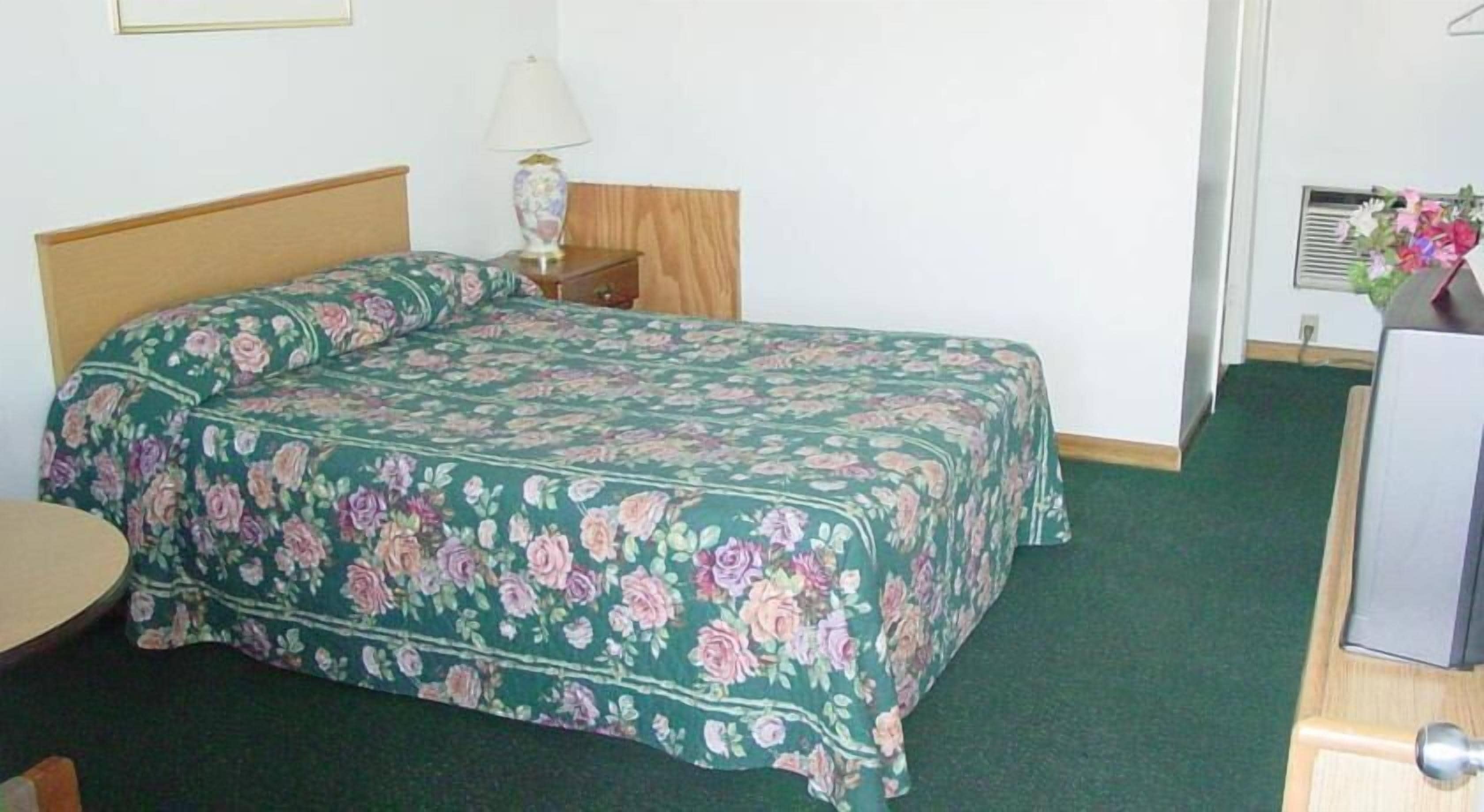 Un letto singolo con copriletto floreale in una stanza con testiera in legno, un comodino con una lampada e un tavolino con una sedia.