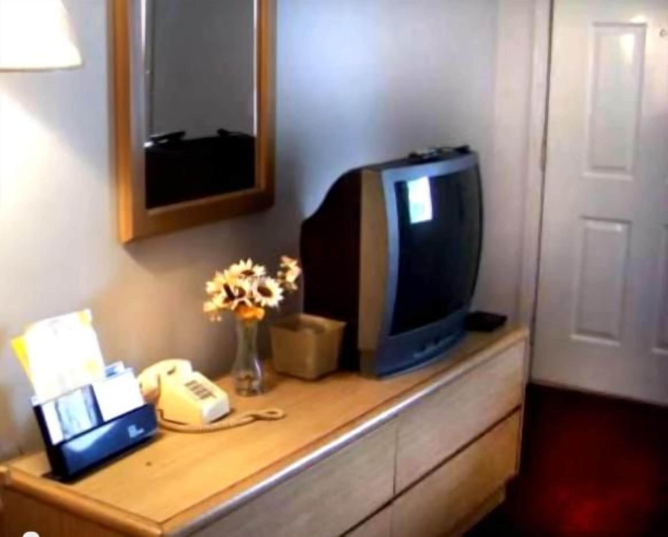 A room with a wooden dresser, a television, a vase with flowers, a telephone, and a lamp.