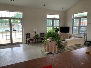 Una habitación amplia con ventanas grandes, televisor, una mesa con una planta y una silla de ruedas.