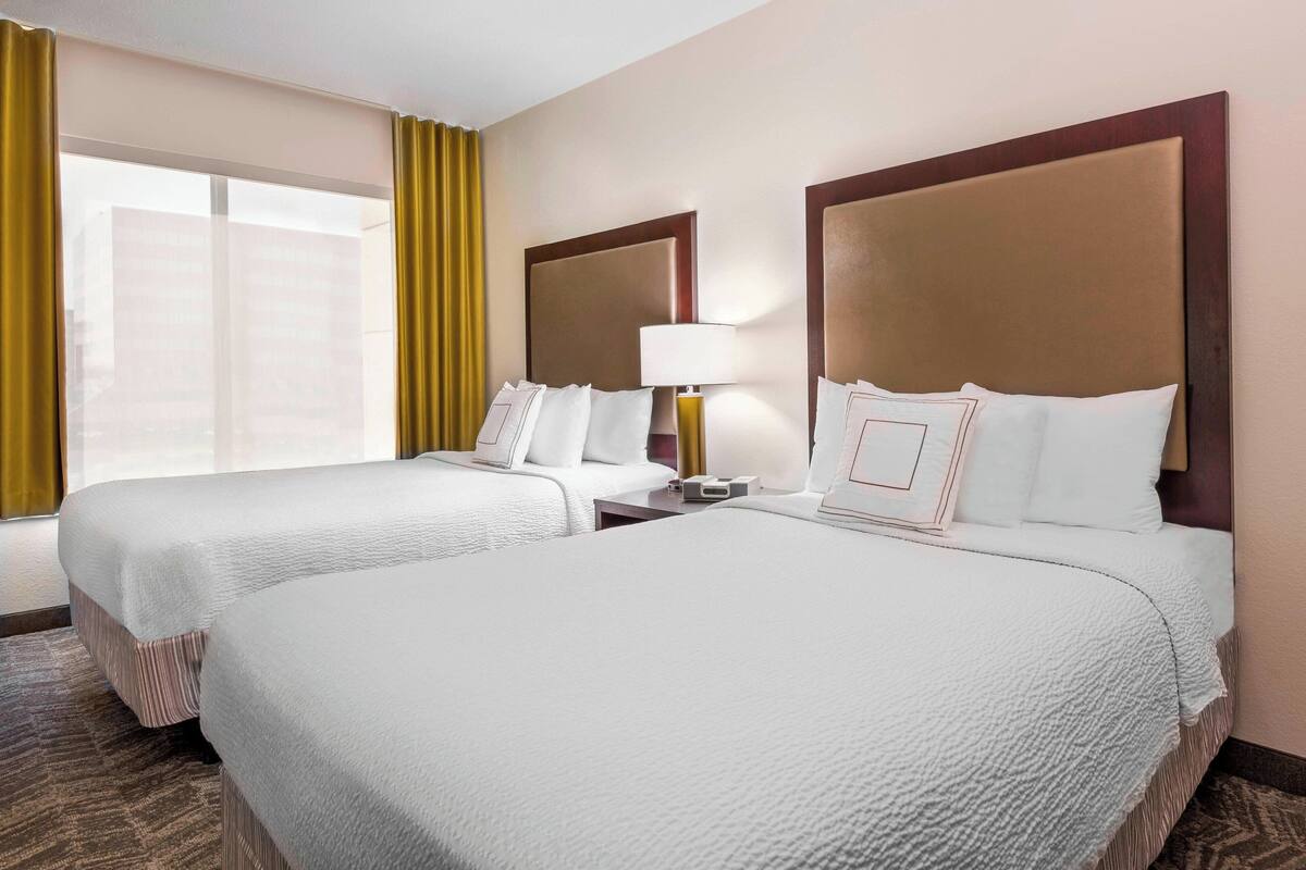 A hotel room with two beds, a nightstand, a lamp, and a window with curtains.