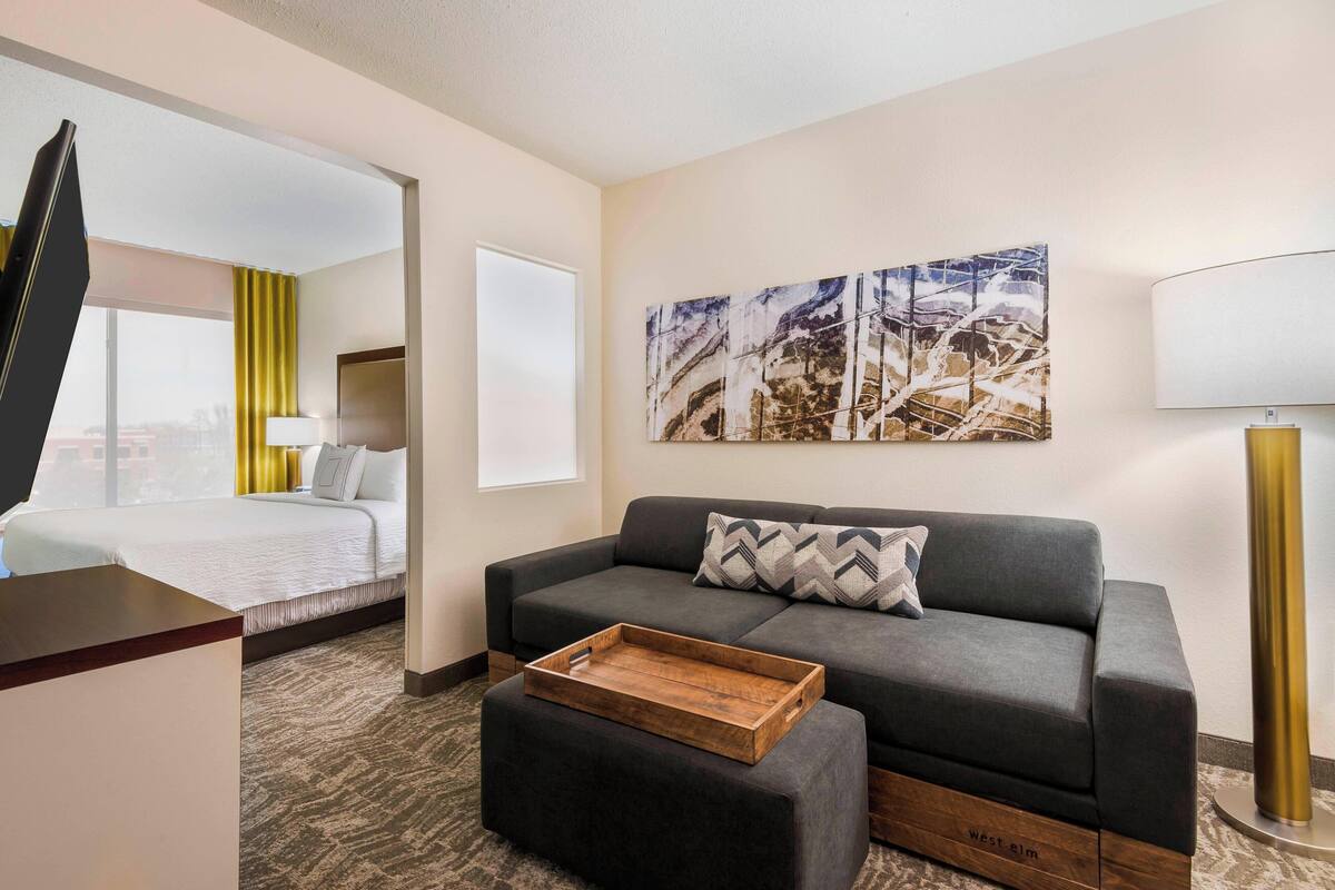 A hotel room with a bed, a gray sofa, a wooden tray, and a large abstract painting on the wall.
