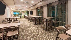Un comedor de hotel con varias mesas y sillas, una alfombra estampada y ventanas grandes con cortinas.