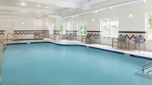 Une piscine dont le bord est recouvert de carreaux, des chaises disposées autour de la piscine et des fenêtres avec des barreaux.