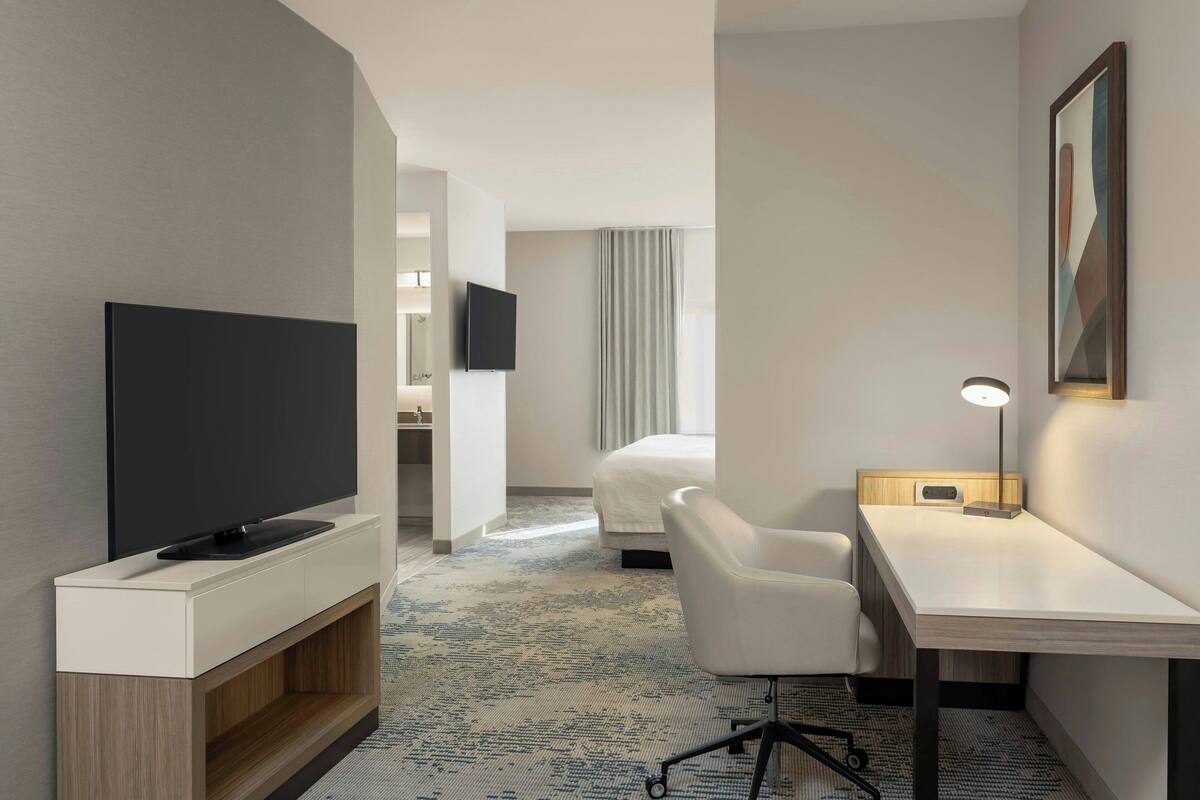 A hotel room with a flat-screen TV on a wooden stand, a desk with a lamp, a white chair, and a bed with a grey curtain.