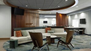 Un hall d’hôtel moderne avec un canapé d’angle, deux fauteuils, une petite table basse, un téléviseur fixé au mur et un coin bar avec des tabourets.