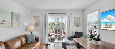 A modern living room with a leather sofa, wooden coffee table, and a dining table with chairs. There is a large window with curtains, a view of the outdoors, and a framed picture on the wall.