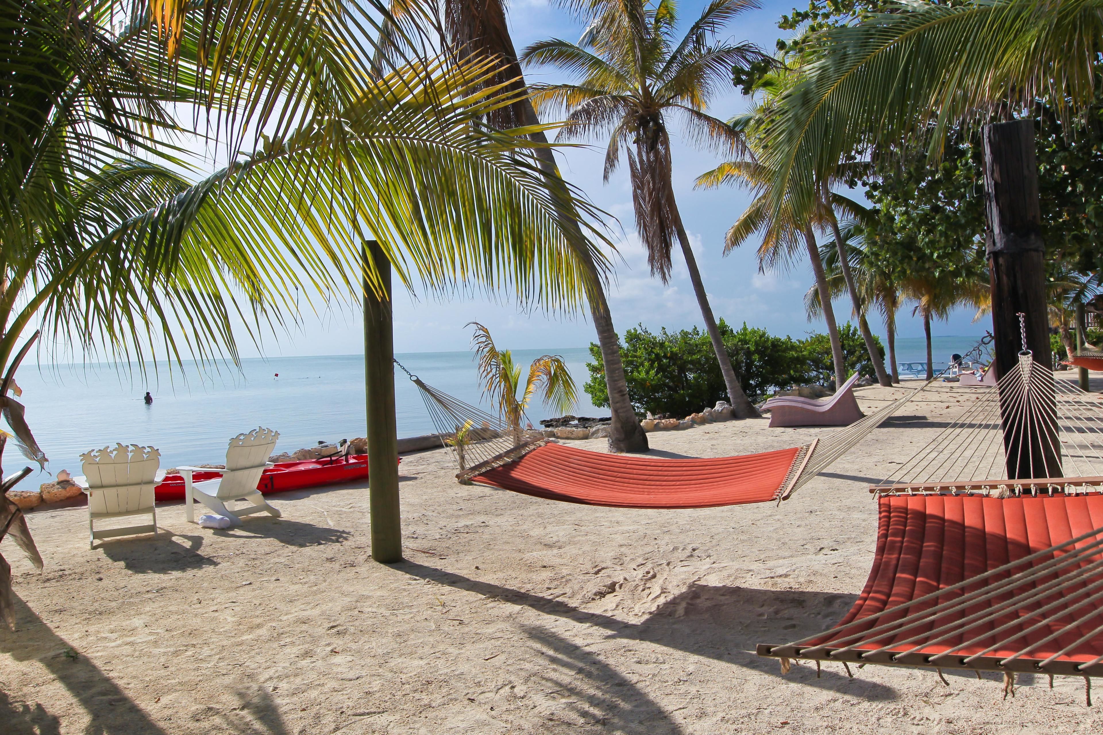 A beach with a hammock, white chairs, and a red lounge chair.