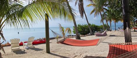 A beach with a hammock, white chairs, and a red lounge chair.