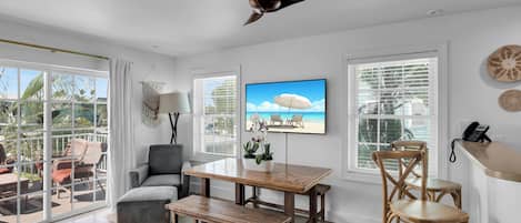 A spacious living area with a wooden dining table, comfortable seating, a ceiling fan, and large windows offering a view of palm trees.