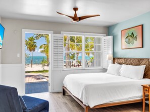 A bedroom with a bed, a ceiling fan, a window with shutters, a framed picture on the wall, and a view of the beach.
