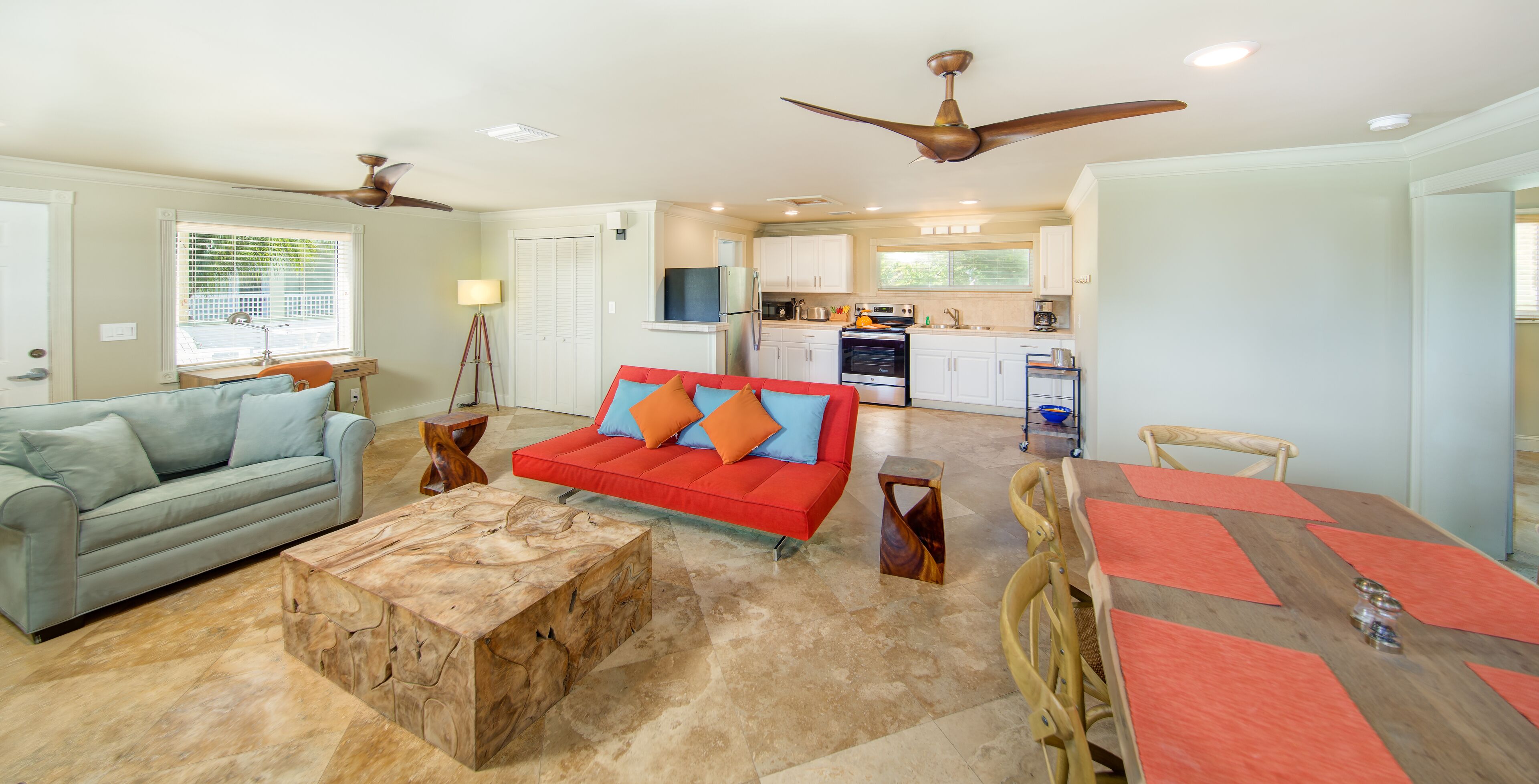 A spacious living room with a red sofa, a grey couch, and a wooden coffee table. There is a dining area with a wooden table and chairs. The kitchen is visible in the background with white cabinets and a microwave.