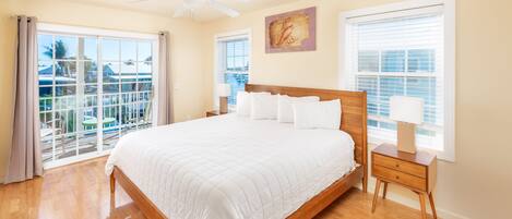 A bedroom with a large bed, a ceiling fan, and a balcony with a view of buildings and palm trees.