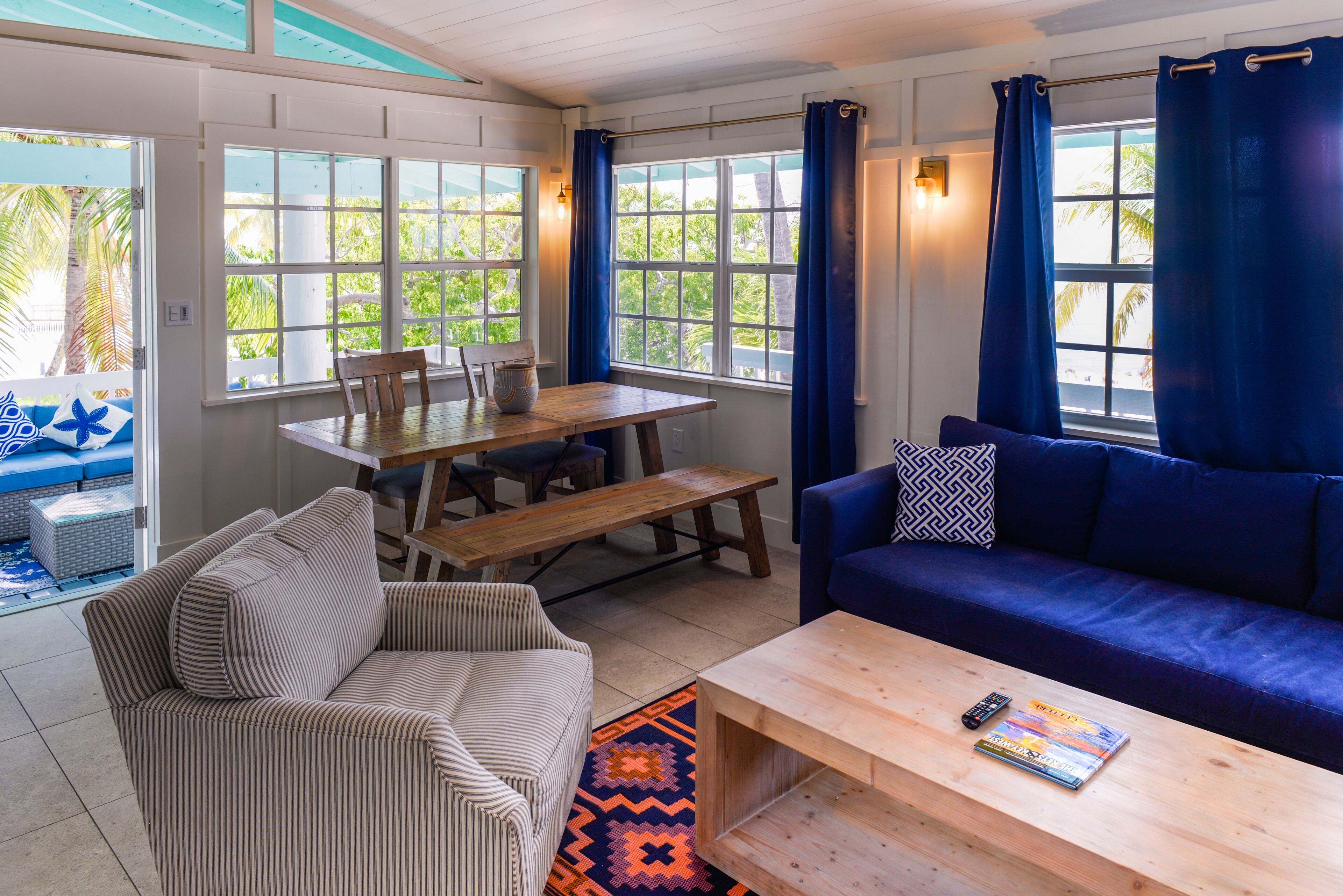 Un salon chaleureux avec une table en bois, un canapé bleu, un divan à rayures et une vue sur l’extérieur grâce à de grandes fenêtres.
