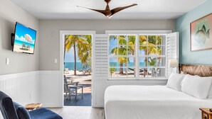 A bedroom with a large bed, a ceiling fan, a TV on the wall, and a view of the beach through a window. - Lime Tree Bay Resort (Long Key)
