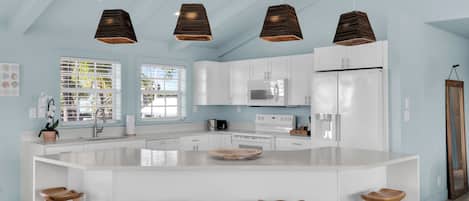 A modern kitchen with white cabinetry, a large island with stools, pendant lighting, and a view of the outdoors through large windows.