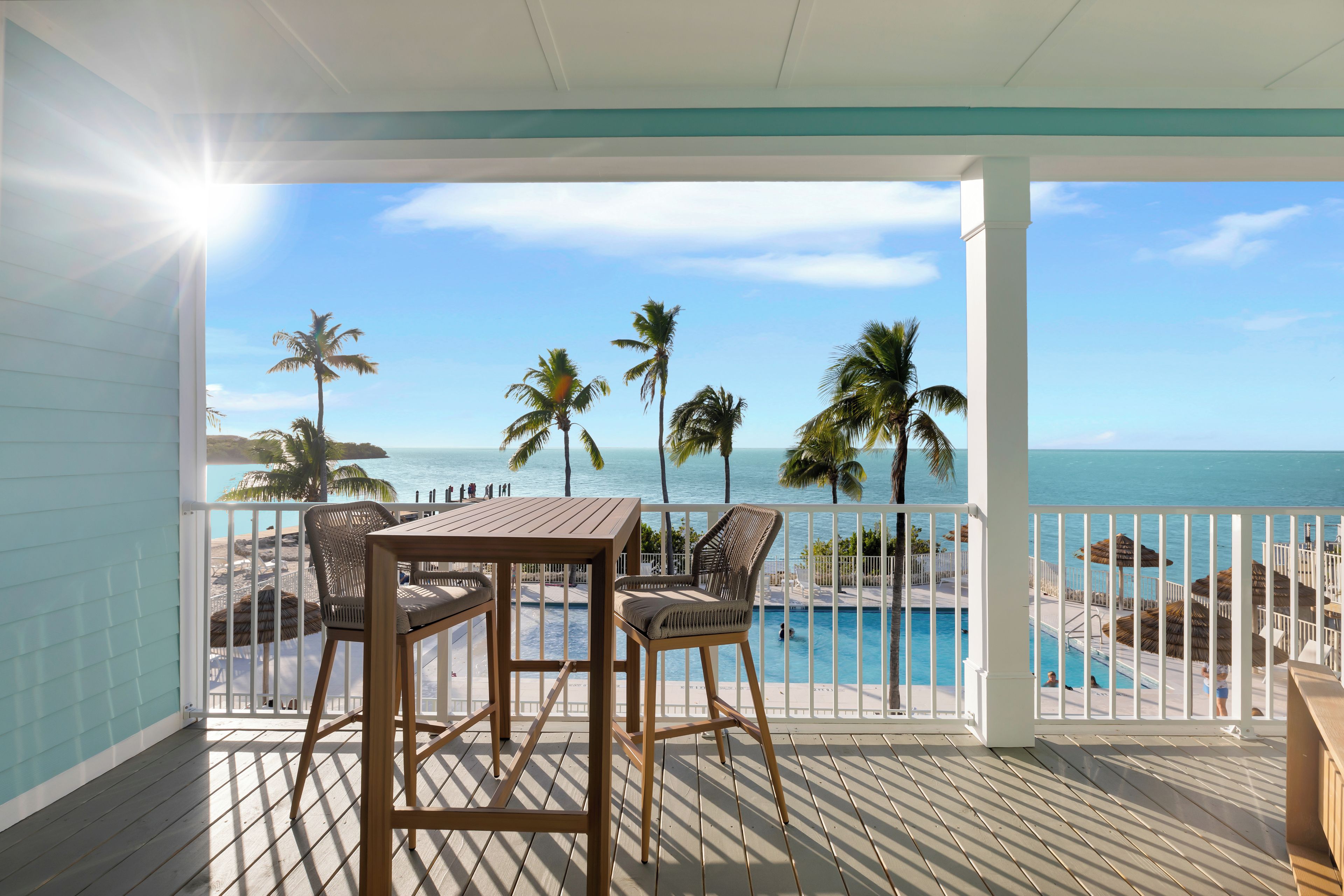 A wooden deck with a table and chairs, overlooking a pool and beach with palm trees.