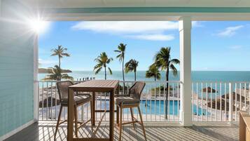 A wooden deck with a table and chairs, overlooking a pool and beach with palm trees.