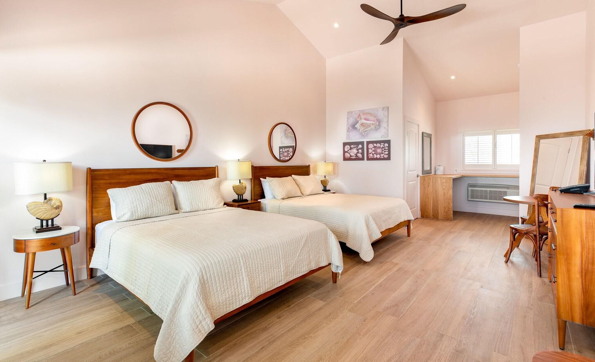 A hotel room with two beds, a ceiling fan, a desk with a chair, and a window with blinds.