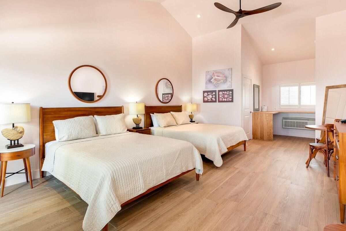 Una habitación de hotel con dos camas, un ventilador de techo, un escritorio con silla y una ventana con persianas.