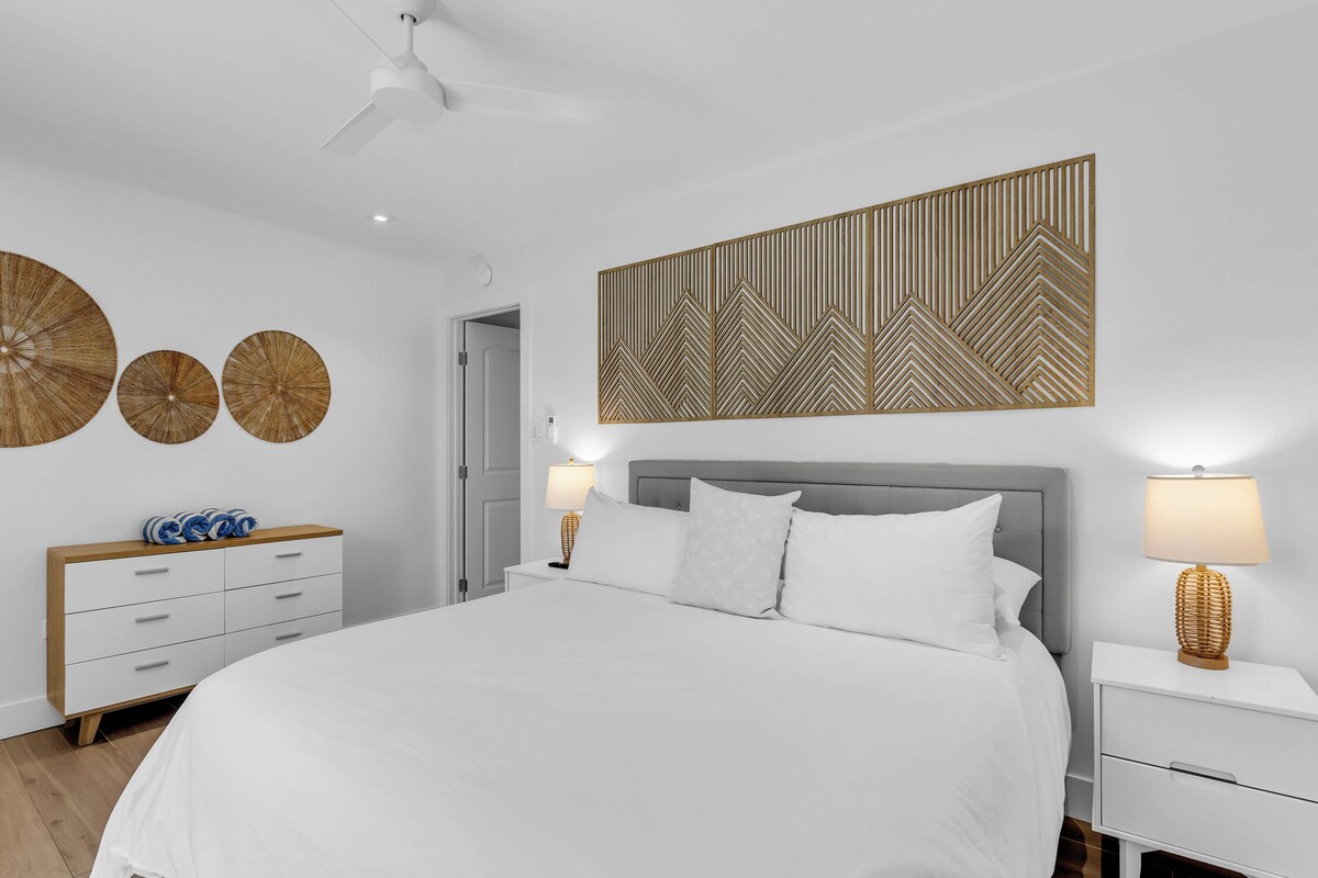 Une chambre moderne avec un grand lit, deux tables de chevet avec des lampes, un ventilateur de plafond et des œuvres d’art décoratives au mur.