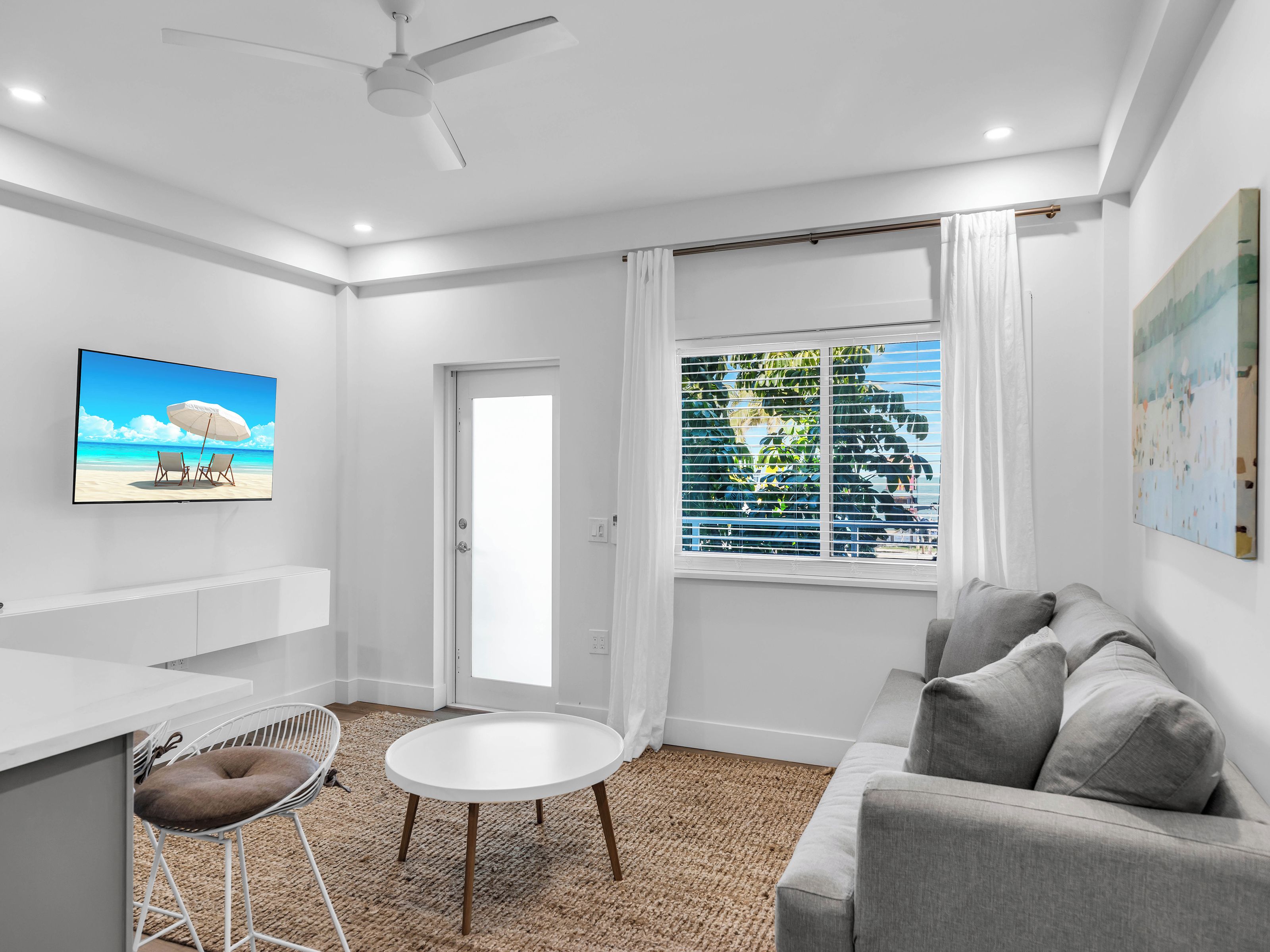 A modern living room with a white sofa, a round table, a TV displaying a beach scene, and a window with blinds.
