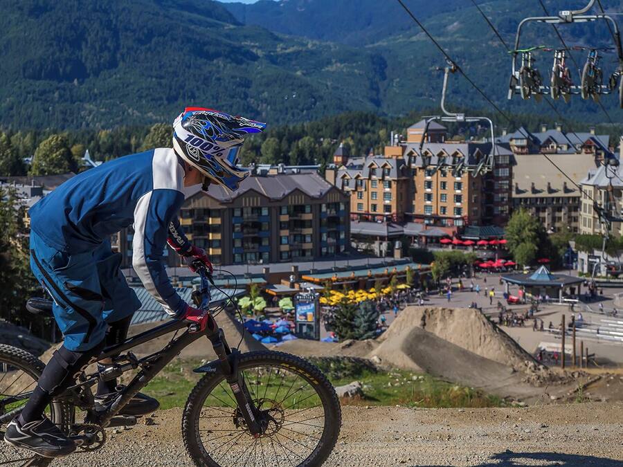 Escena de ciclismo de montaña con un ciclista, un telesilla al fondo y edificios en la distancia.