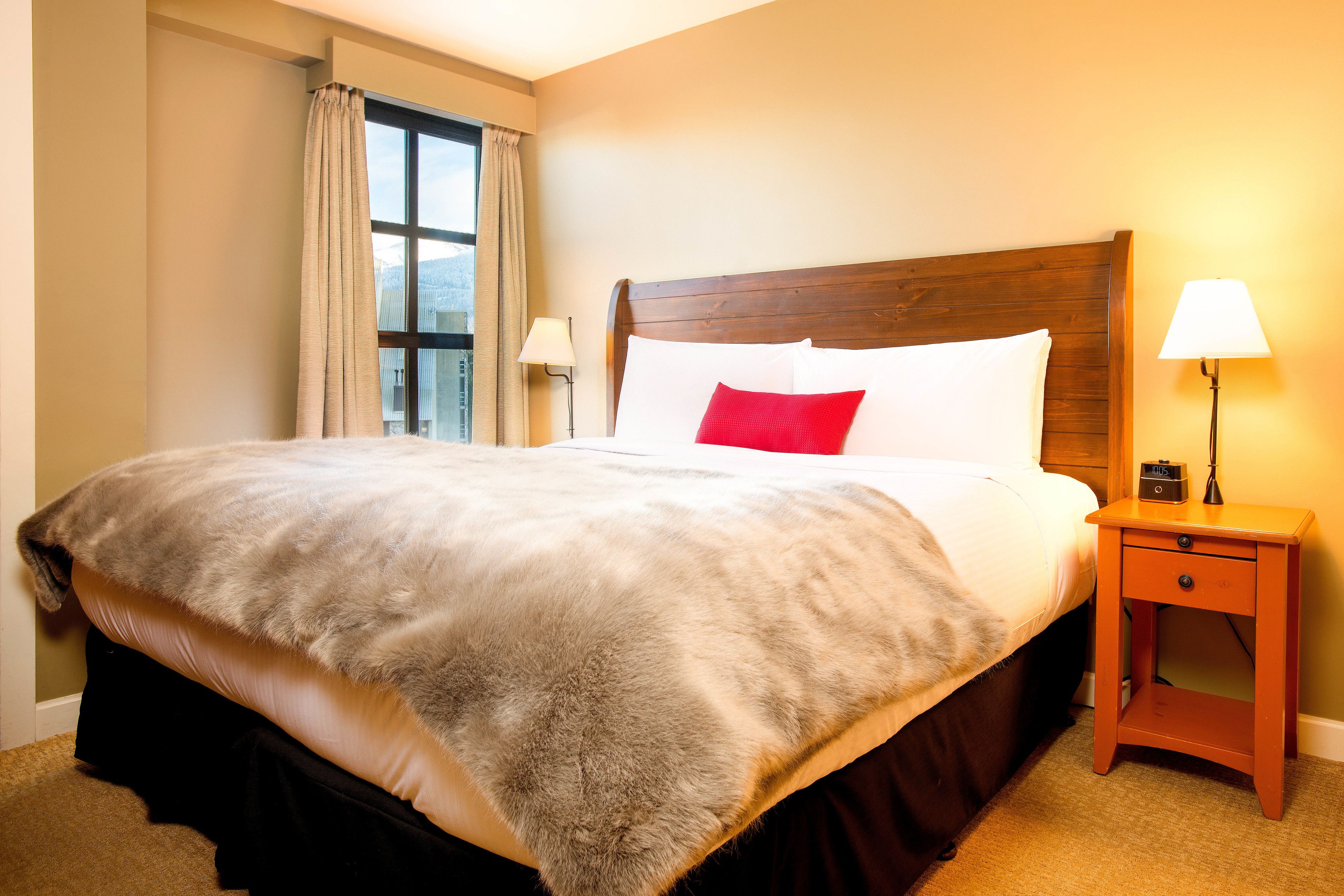 A neatly made bed with a wooden headboard, a bedside table with a lamp, and a window with curtains.